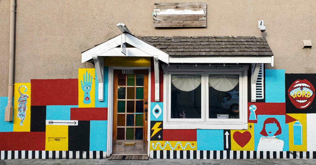Colorful mural on a building exterior featuring geometric patterns, images of hands, hearts, and whimsical figures, with a distinct entrance door.